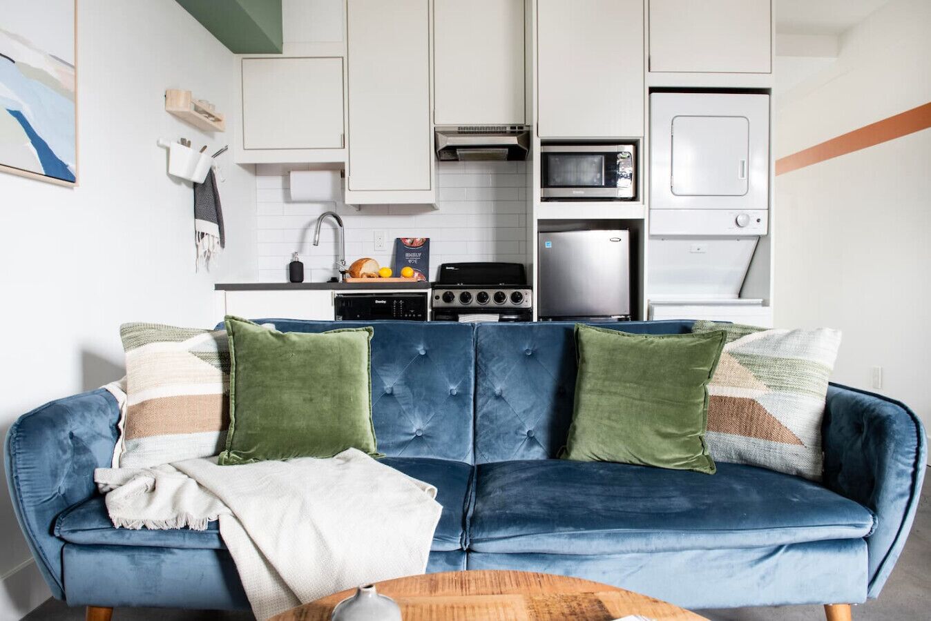 A modern kitchen with white cabinets, a microwave, and a stove. There is a blue sofa with green and beige cushions in the living area.