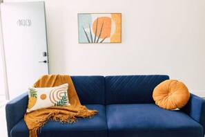 A blue sofa with a mustard throw blanket and two pillows, one with a leaf pattern and one round. A framed abstract painting hangs on the wall above.