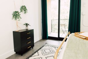Une chambre à coucher avec un lit, une commode noire, une plante suspendue et une vue sur l’extérieur grâce à une porte coulissante.