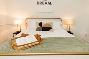 A neatly made bed with a green blanket, a wicker basket with an open book, and two bedside lamps on either side.