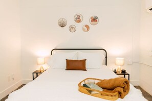 A neatly made bed with white linens and a brown pillow, flanked by two bedside tables with lamps, a wall-mounted artwork, and a wicker basket with a book and a glass on the bed.