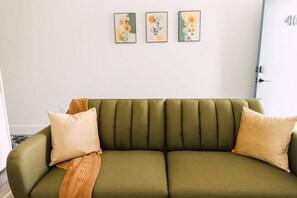 A green sofa with two beige pillows and a brown throw blanket. Three framed floral artworks are hung on the wall above the sofa.