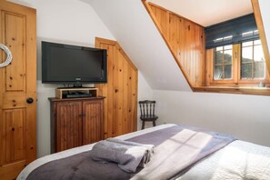 A bedroom with a sloped ceiling, a television mounted on a wooden cabinet, a bed with a blanket, and a window with a view.