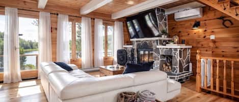 Interior acogedor de una cabaña con un sofá blanco grande, una chimenea de piedra y un televisor montado en la pared.