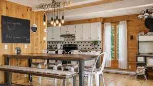 A rustic wooden interior with a dining area, white chairs, a wooden bench, a chalkboard with text, and a kitchen with white cabinets and a stove. - Le Martini Bord de l eau, spa, piscine. (Entrelacs)