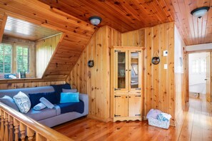 A wooden cabin interior with a sloped ceiling, a sofa with cushions, a cabinet, and a basket on the floor.