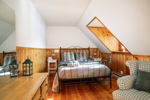 A bedroom with a sloped ceiling, wooden paneling, a metal bed frame, a bedside table, a lamp, and a checkered chair.