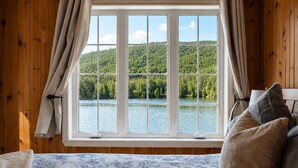 Una habitación con un ventanal grande, paredes de madera, una cama con colcha estampada y vistas a un lago y un bosque.