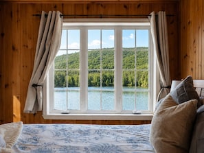 Une pièce dotée d’une grande fenêtre, de murs en bois, d’un lit recouvert d’une couvre-lit à motifs floraux, et offrant une vue sur un lac et une forêt.
