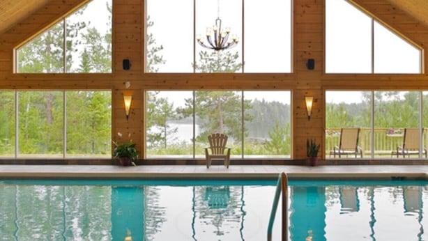 Un espace piscine avec des murs en bois et de grandes fenêtres, un lustre et un fauteuil placé au bord de la piscine.