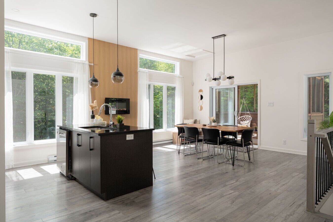 Une cuisine moderne dotée d’un îlot central, d’un coin repas avec des chaises, de grandes fenêtres et d’un escalier.