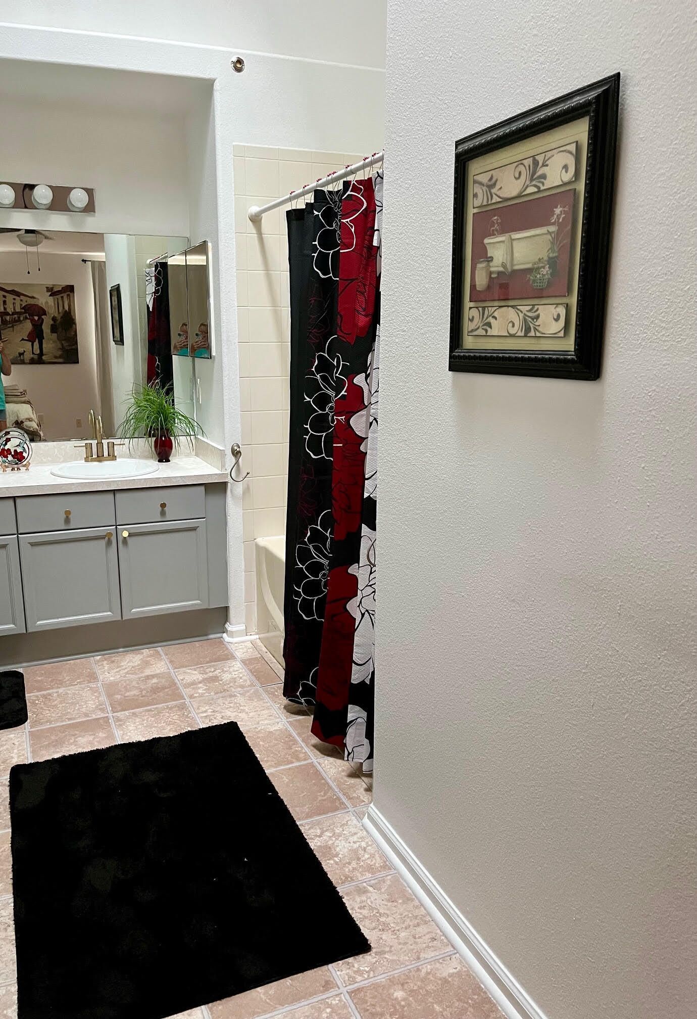 Une salle de bain avec un lavabo blanc, des meubles gris, un tapis noir, un rideau de douche à motifs floraux et un tableau encadré au mur.