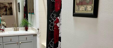 Une salle de bain avec un lavabo blanc, des meubles gris, un tapis noir, un rideau de douche à motifs floraux et un tableau encadré au mur.