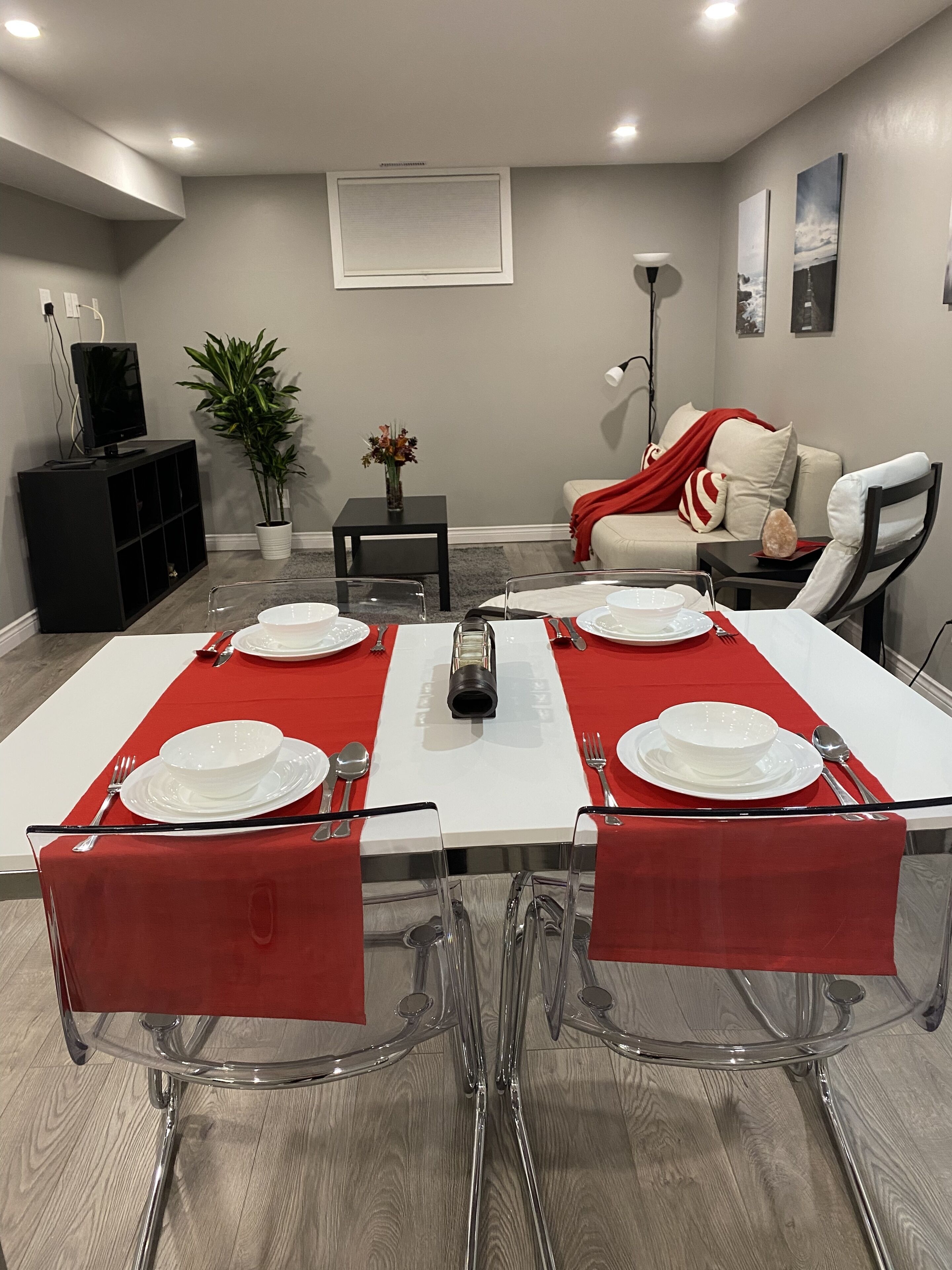 A dining area with a rectangular table set for four, surrounded by clear glass chairs with red cushions.