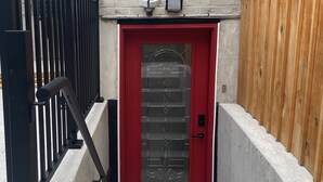 Une porte rouge avec un panneau en verre, un portail en métal et un escalier en béton menant à un bâtiment à la façade en briques.