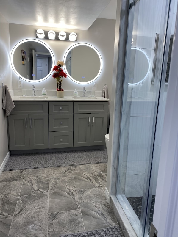 Un baño moderno con tocador, dos espejos con luz, zona de ducha y suelo de mármol.