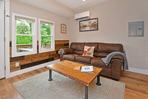 A living room with a brown leather sofa, a wooden coffee table, and a view of greenery through large windows. - Pet Friendly - Close to Town - Deck & Fire Pit (Lake George)