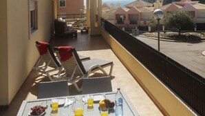 Varanda com móveis de vime, uma mesa com frutas e bebidas, e vista para edifícios e uma colina.
