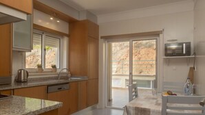 Cozinha moderna com bancadas de granito, armários de madeira e lava-louças embutida. Há uma área de jantar com mesa e cadeiras, e uma grande porta de vidro deslizante que dá para uma varanda com vista para uma paisagem rochosa.
