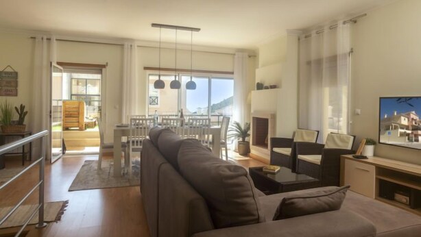 Uma sala de estar espaçosa com um sofá grande, uma área de jantar com mesa e cadeiras, uma lareira e uma janela ampla com vista para o exterior.