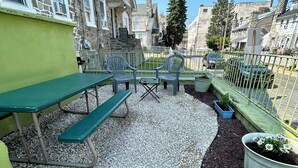 Eine Terrasse mit grünem Tisch und Bänken, umgeben von Kies und Mulch, mit einem Metallzaun und Blumentöpfen.