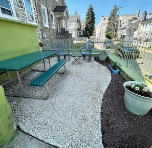 A patio area with a green table and benches, surrounded by gravel and mulch, with a metal fence and potted plants.