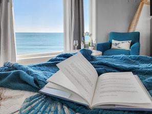 Una habitación con vista al mar, una silla azul y un libro abierto sobre la cama.
