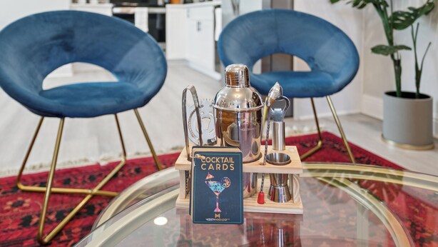 Una carta de cócteles sobre una mesa de vidrio, con un tapete rojo estampado, dos sillas azules y una planta en maceta en un ambiente de cocina moderno.