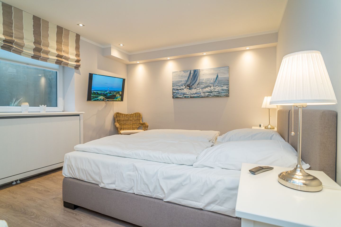 A modern bedroom with a large bed, a bedside table with a lamp, a wicker chair, a TV, and a painting of a sailboat.