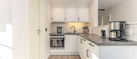 Una cocina moderna con gabinetes blancos, horno empotrado, cafetera y salpicadero de azulejos.