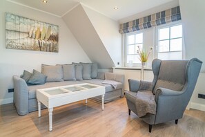 Una sala de estar moderna con un sofá gris, una mesa de centro blanca y un sillón gris. Hay un ventanal con persianas y un cuadro en la pared.
