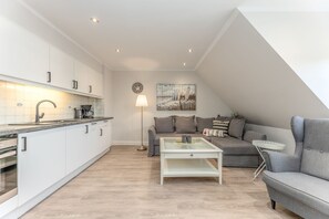 Une cuisine moderne avec des meubles blancs et un four encastré, un canapé gris avec des coussins, une petite table basse et un lampadaire.