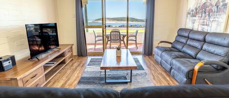 A living room with a sofa, a TV, a wooden table, and a view of the beach through large windows.