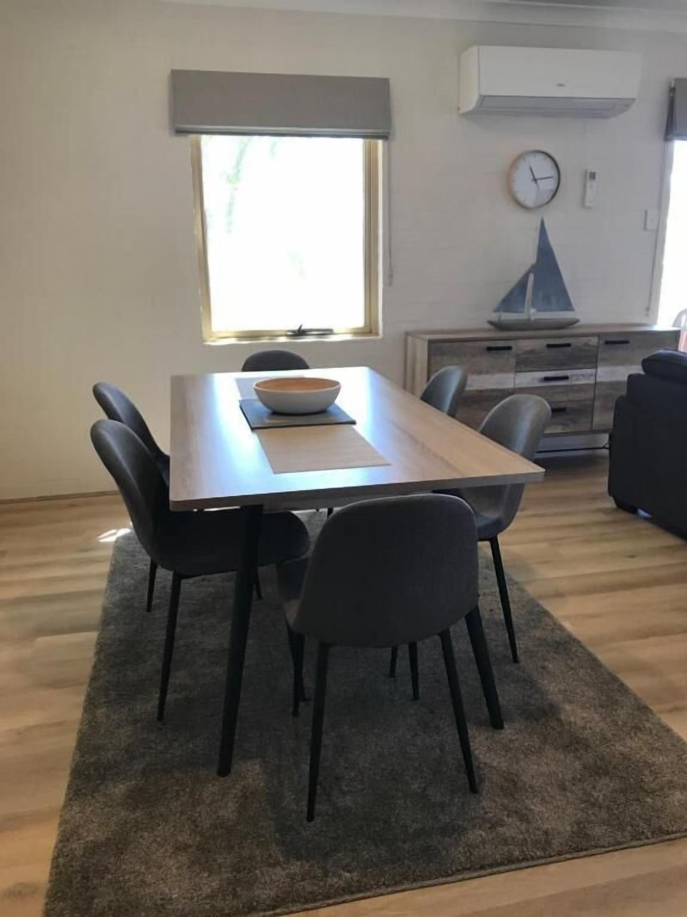 Un coin repas avec une table, des chaises et un saladier. Il y a une fenêtre avec vue, une horloge et un meuble avec des tiroirs.