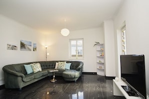 A living room with a sectional sofa, a small round table, a floor lamp, a bookshelf, and a TV. - Ferienwohnung Cosmo - Direkt am Waldrand mit Balkon und in Strandnähe - Ferienwohnung Cosmo (Glowe)