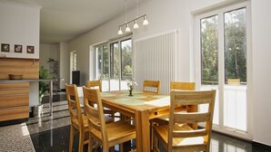 Een ruime eetruimte met een houten tafel en stoelen, grote ramen en een moderne keuken op de achtergrond.