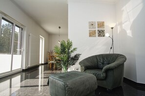 A modern living room with a green armchair, a coffee table, and a potted plant. There is a dining area with wooden chairs and a table in the background. - Ferienwohnung Cosmo - Direkt am Waldrand mit Balkon und in Strandnähe - Ferienwohnung Cosmo (Glowe)