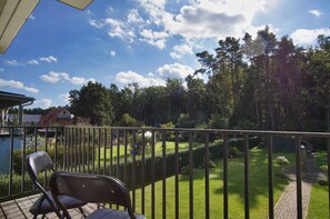 A balcony with a view of a well-maintained lawn and trees, a chair, and a house in the background. - Ferienwohnung Cosmo - Direkt am Waldrand mit Balkon und in Strandnähe - Ferienwohnung Cosmo (Glowe)