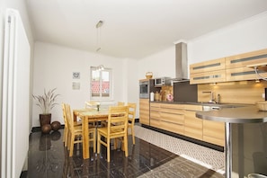 A modern kitchen with wooden cabinets, a dining area with a table and chairs, and a window with a view of a red-roofed building. - Ferienwohnung Cosmo - Direkt am Waldrand mit Balkon und in Strandnähe - Ferienwohnung Cosmo (Glowe)