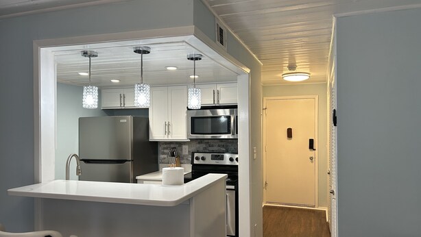 A compact kitchen with white cabinets, a stainless steel refrigerator, a microwave, and an oven. There are two pendant lights above the island. - The Dolce Cabana located on the golf course in beautiful Diamondhead, MS. (Diamondhead)