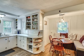 A kitchen with white cabinetry, a dining area with wooden chairs, and a window with blinds. - Near Beach: Coastal Maine Getaway w/ Yard (Wells)