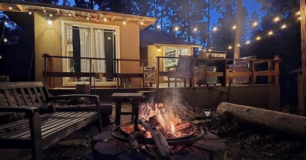A wooden deck with a fire pit, outdoor seating, and a building with string lights.