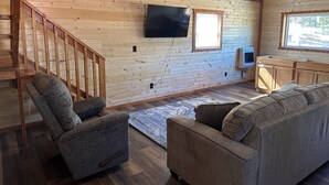 A cozy cabin interior with a wooden staircase, a flat-screen TV mounted on the wall, a small kitchen area with cabinets, and a large window providing natural light. - White cabin in the woods close to Univ. of Central Missouri and Whiteman AF Base (Warrensburg)