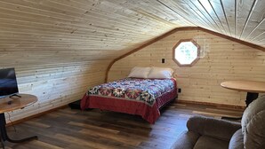 A cozy attic bedroom with a wooden sloped ceiling, a bed with a colorful quilt, a small round table, and a comfortable chair. - White cabin in the woods close to Univ. of Central Missouri and Whiteman AF Base (Warrensburg)