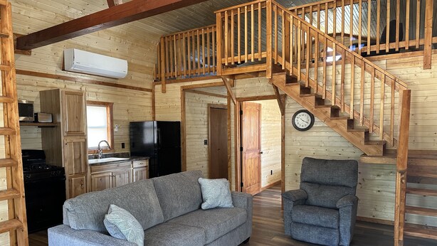 A cozy cabin interior with a wooden staircase, a sofa, a chair, a kitchen area, and a clock on the wall. - White cabin in the woods close to Univ. of Central Missouri and Whiteman AF Base (Warrensburg)