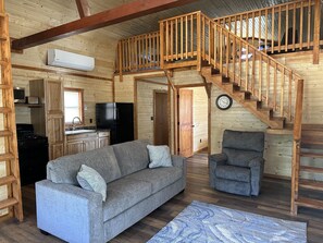 L’intérieur d’un chalet accueillant, avec un escalier en bois, un canapé, un fauteuil, un coin cuisine et une horloge au mur.