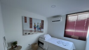Une petite chambre propre, avec un lit simple, un climatiseur fixé au mur, une fenêtre avec des stores, une petite table sur laquelle est posée une plante, et un tableau accroché au mur.
