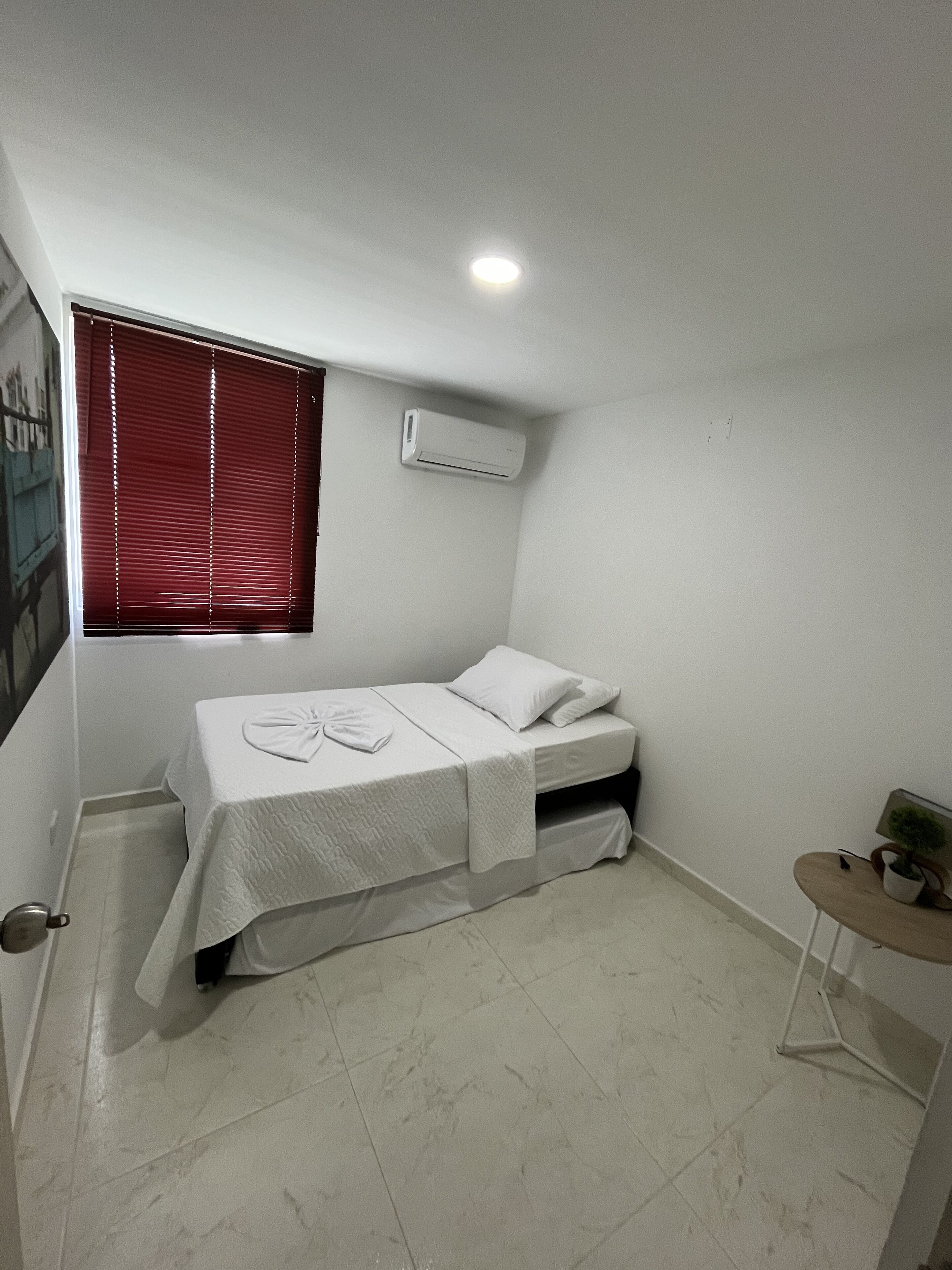 Une petite pièce propre, avec un lit simple, une petite table et une fenêtre avec des stores.