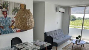 Un salon moderne avec un canapé gris, une table à manger en verre sur laquelle sont posés des journaux, et un grand luminaire en fibres tressées.