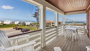 A covered porch with white railings and a wooden deck, overlooking a beach and a road with parked cars. - Endless Summer: 1-Min Walk Beach, Pool, Hot Tub (Kitty Hawk)